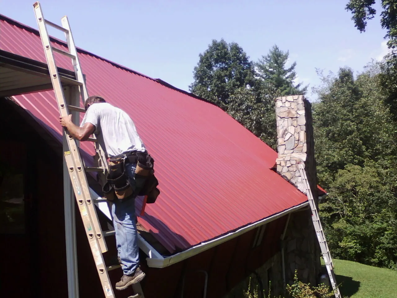Roofer on a ladder beside a red metal roof and stone chimney — Soffit & Fascia Repair by Roofing Brothers in Anita